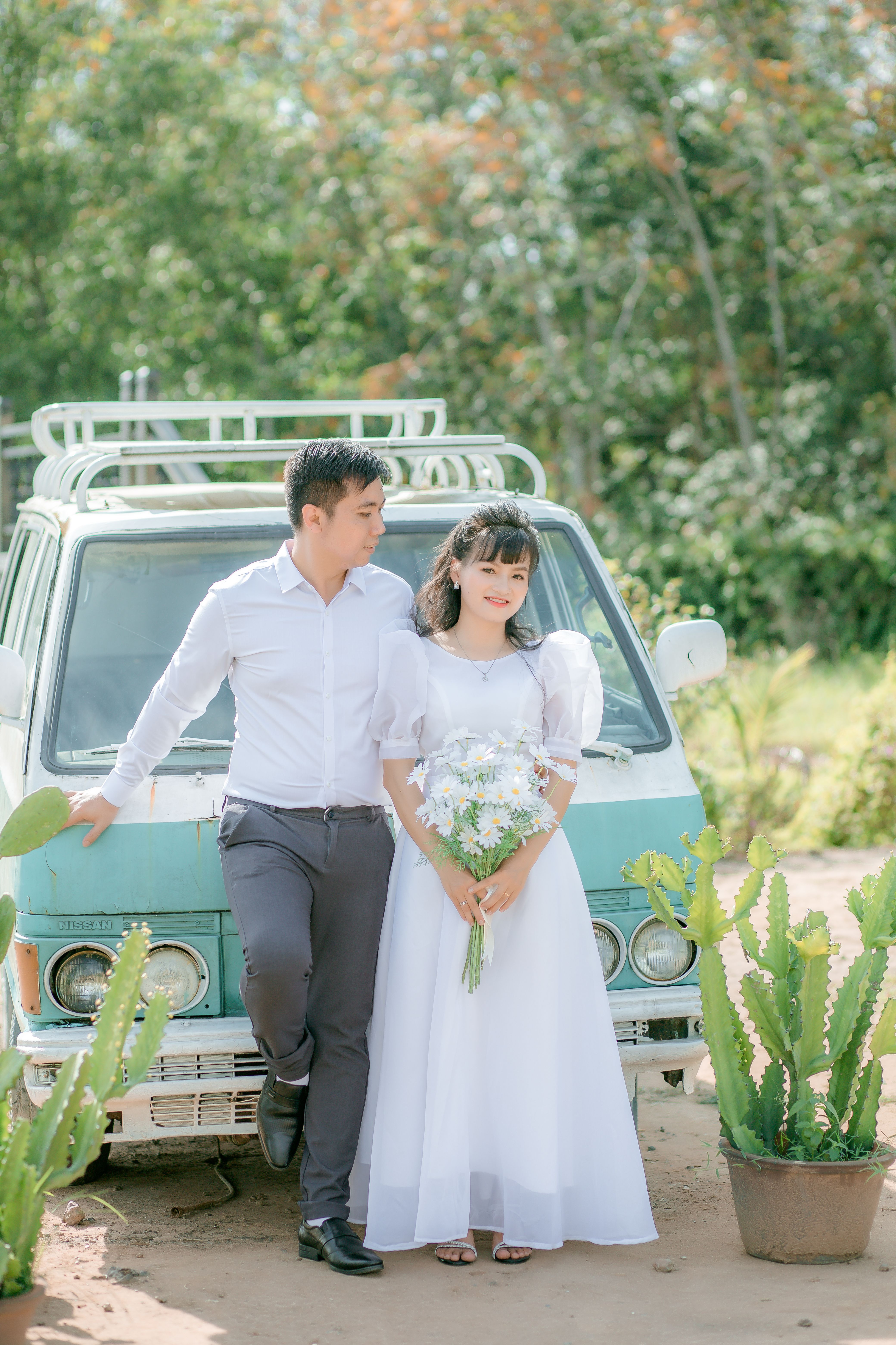 A couple shares a romantic moment outdoors with a bouquet, leaning on a vintage van, captured in bright daylight.