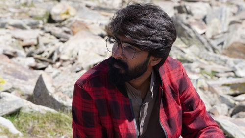 Free Bearded man wearing glasses and plaid jacket, seated outdoors on rocky terrain in Chilās. Stock Photo