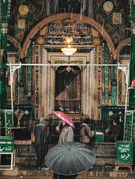 People with umbrellas walk through rain in front of a decorated historical building in Srinagar.