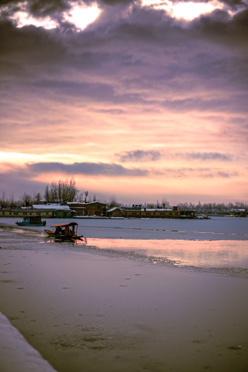 Free A peaceful winter sunset reflecting on a frozen lake with a rustic boat and distant village visible. Stock Photo
