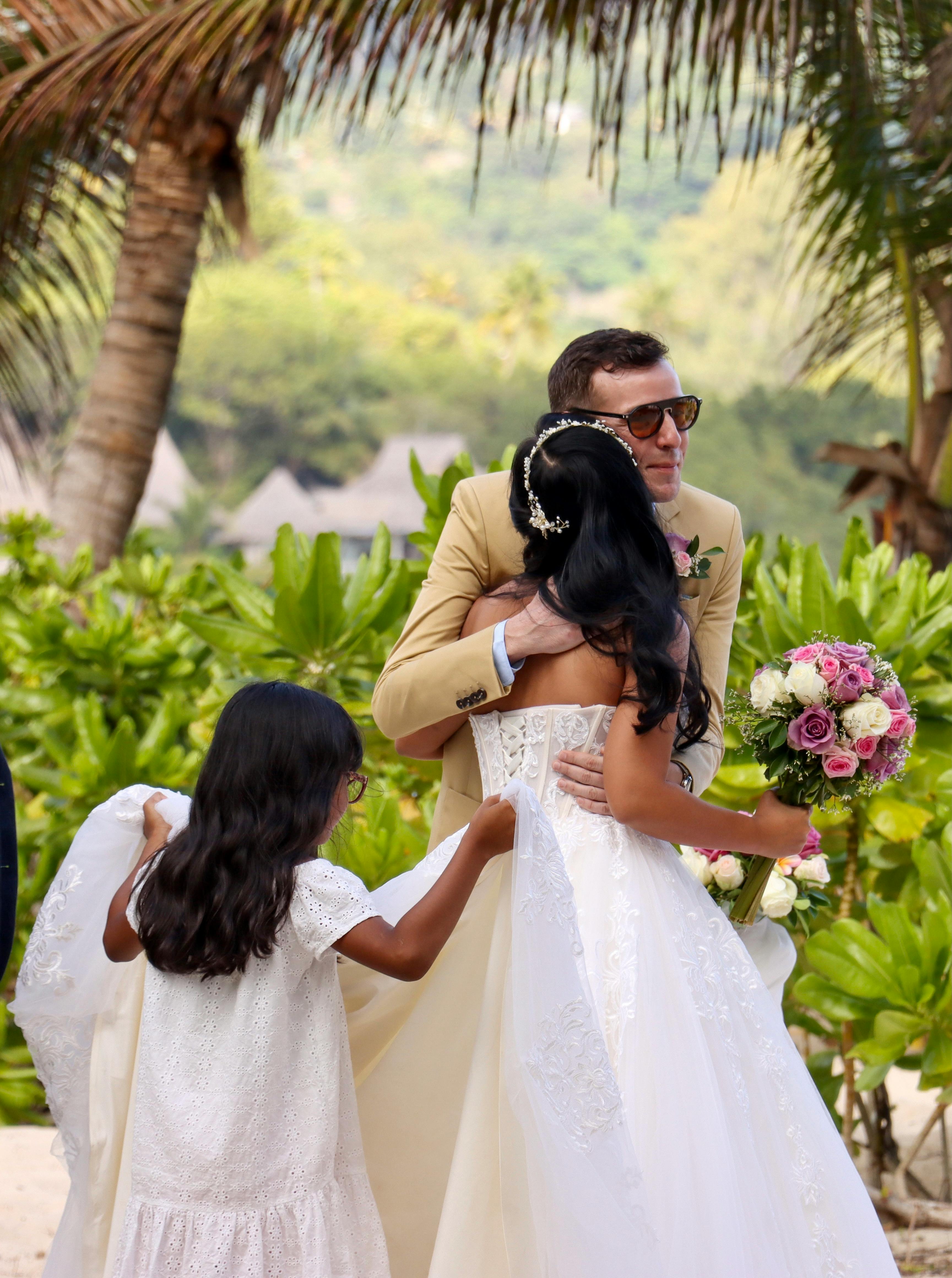 Bride and groom embrace outdoors, with a child holding the bridal gown.