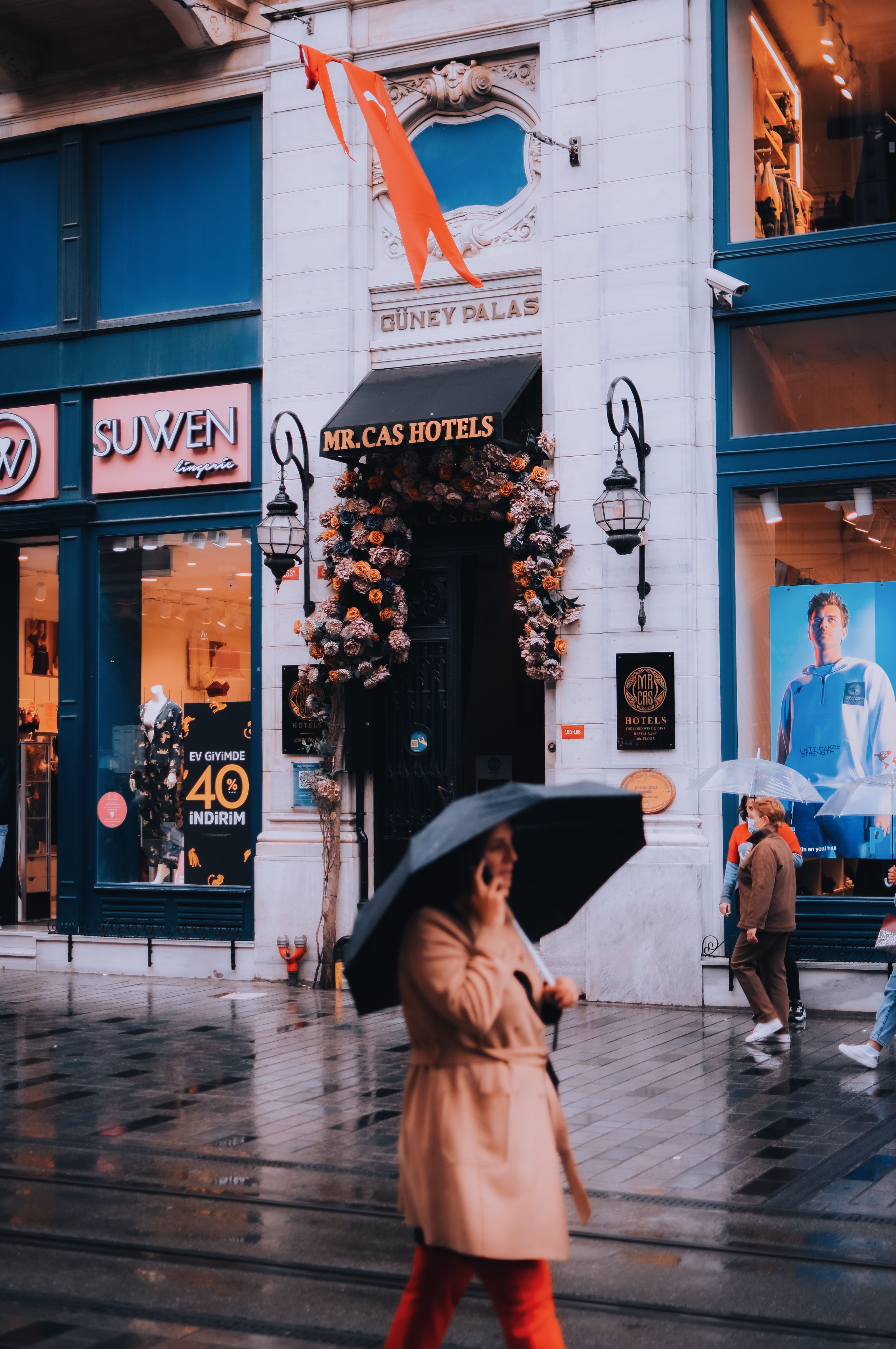 A bustling street scene in Istanbul with people walking under umbrellas past shops and hotels in the rain.