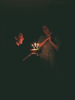 A man presents a lit birthday cake to a woman in a dark room, creating a cozy atmosphere.