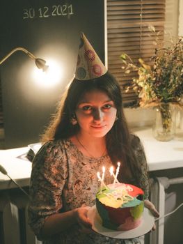 Woman wearing party hat holding a colorful cake with candles indoors.