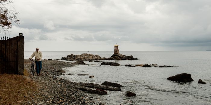 A man walks his bicycle along the rocky seashore of Milazzo, Sicily under a cloudy sky.