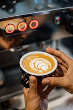 Hands delicately holding a latte with intricate art in a coffee shop setting.