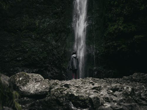 Free Person in Black Jacket Standing on Rocky Ground Near Waterfalls Stock Photo