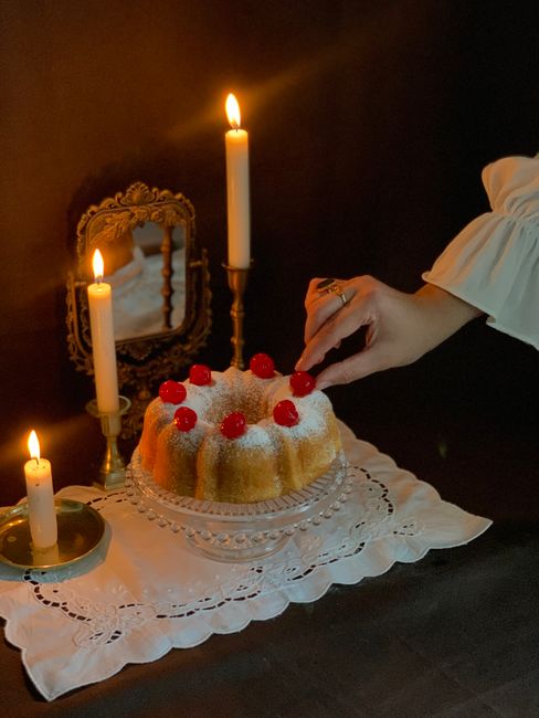 A hand decorates a pound cake with cherries, surrounded by candles and a mirror, in a dimly lit setting.