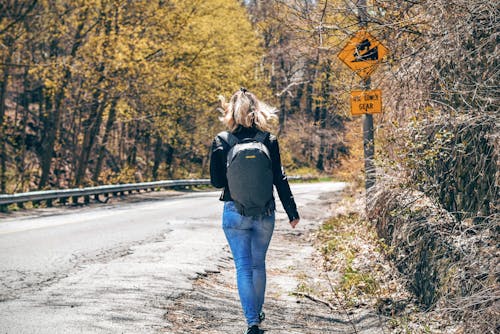 Woman in Blue Denim Fitted Jeans and Wearing Grey Backpack Walking on Gray Asphalt Road Near Road Signage and Trees at Daytime