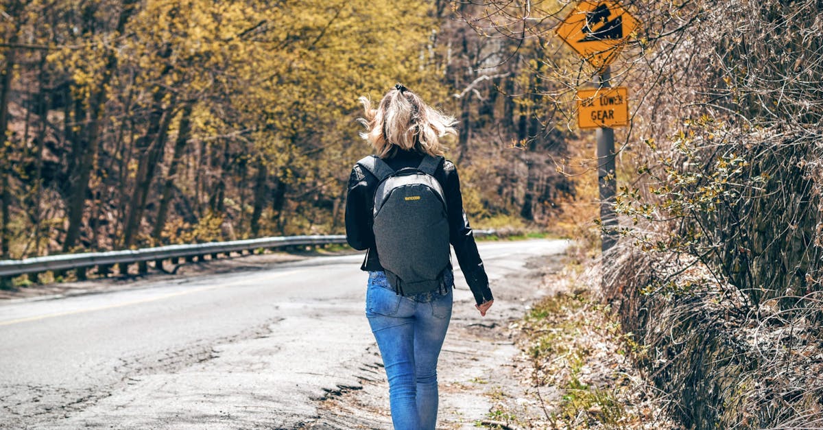Woman in Blue Denim Fitted Jeans and Wearing Grey Backpack Walking on Gray Asphalt Road Near Road Signage and Trees at Daytime