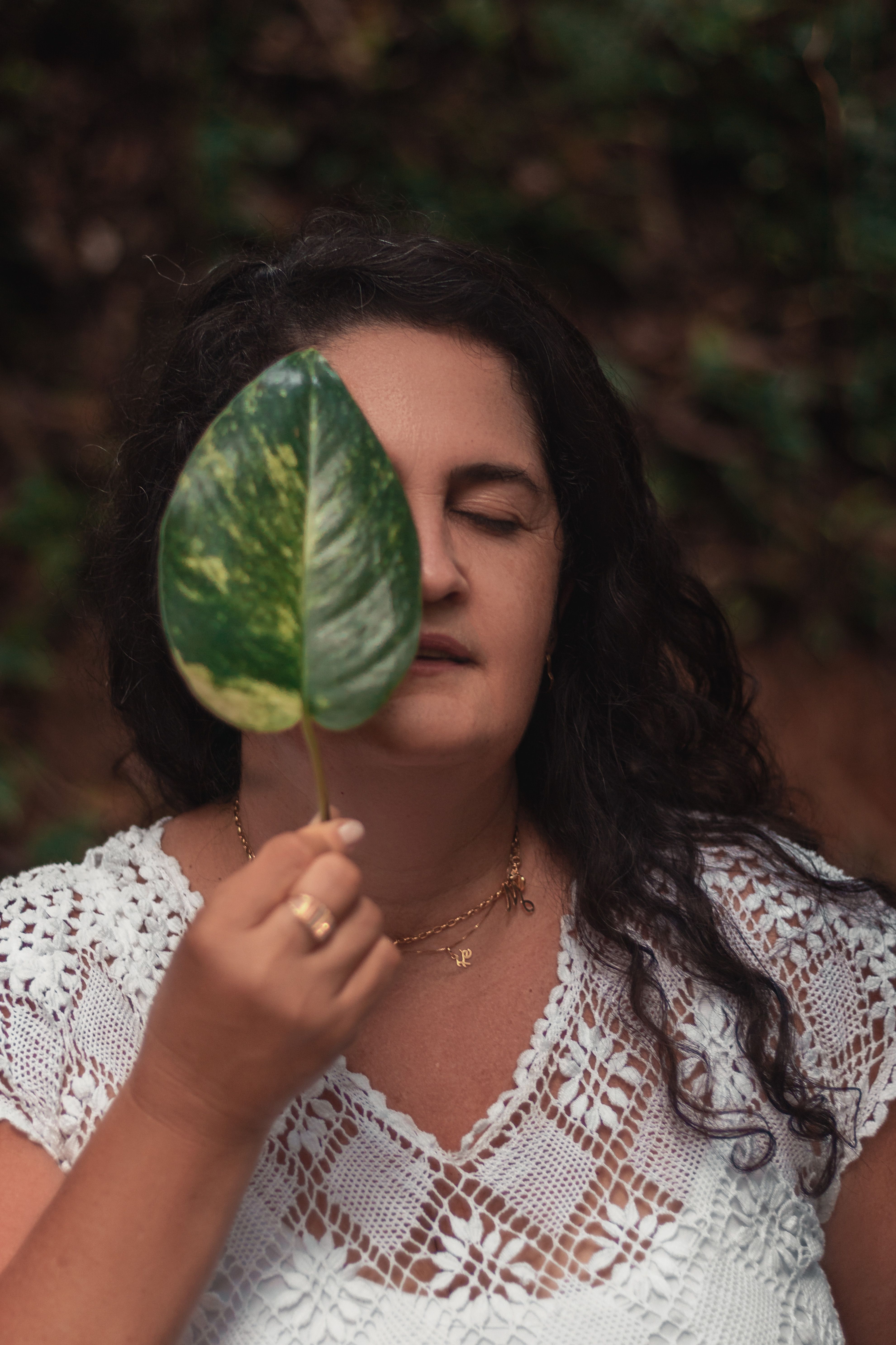 Woman in a white dress holds a leaf over her eye, blending with natural surroundings.
