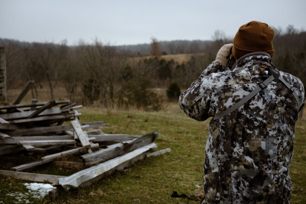Chasseur vu de dos levant des jumelles pour observer une clairière herbeuse derrière une clôture en bois