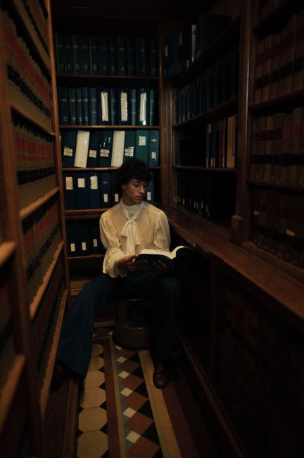 A young man in a retro outfit reading a book in a vintage library, evoking elegance and knowledge.
