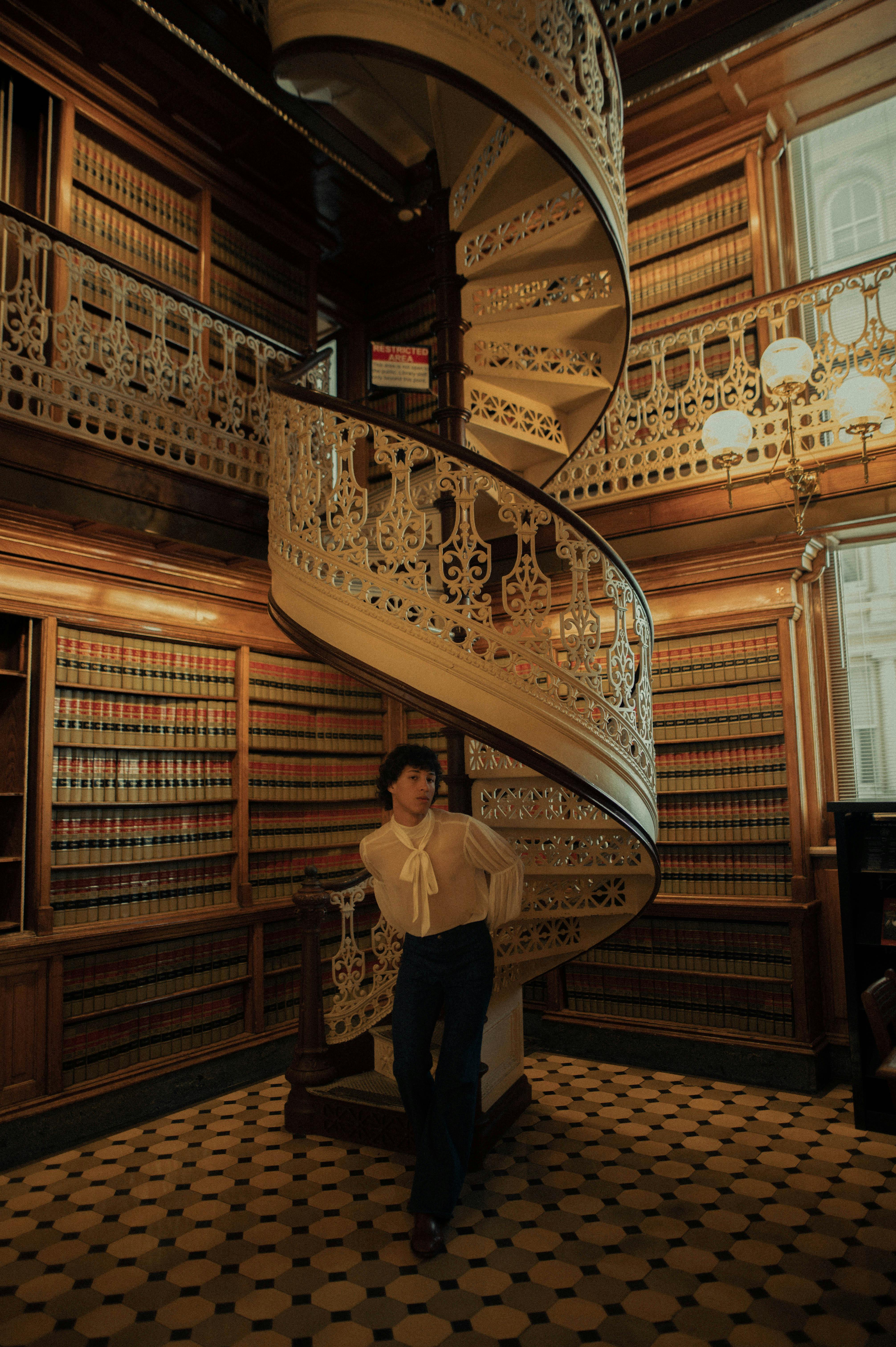A person poses near an ornate spiral staircase in a vintage library.