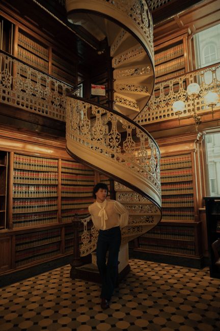A person poses near an ornate spiral staircase in a vintage library.