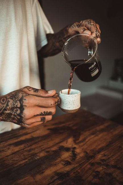 A tattooed barista pouring freshly brewed coffee into a ceramic cup on a wooden table.