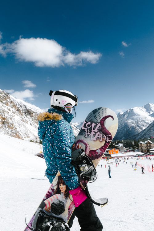 Free A snowboarder in a blue jacket carries a snowboard against a snowy mountain backdrop. Stock Photo