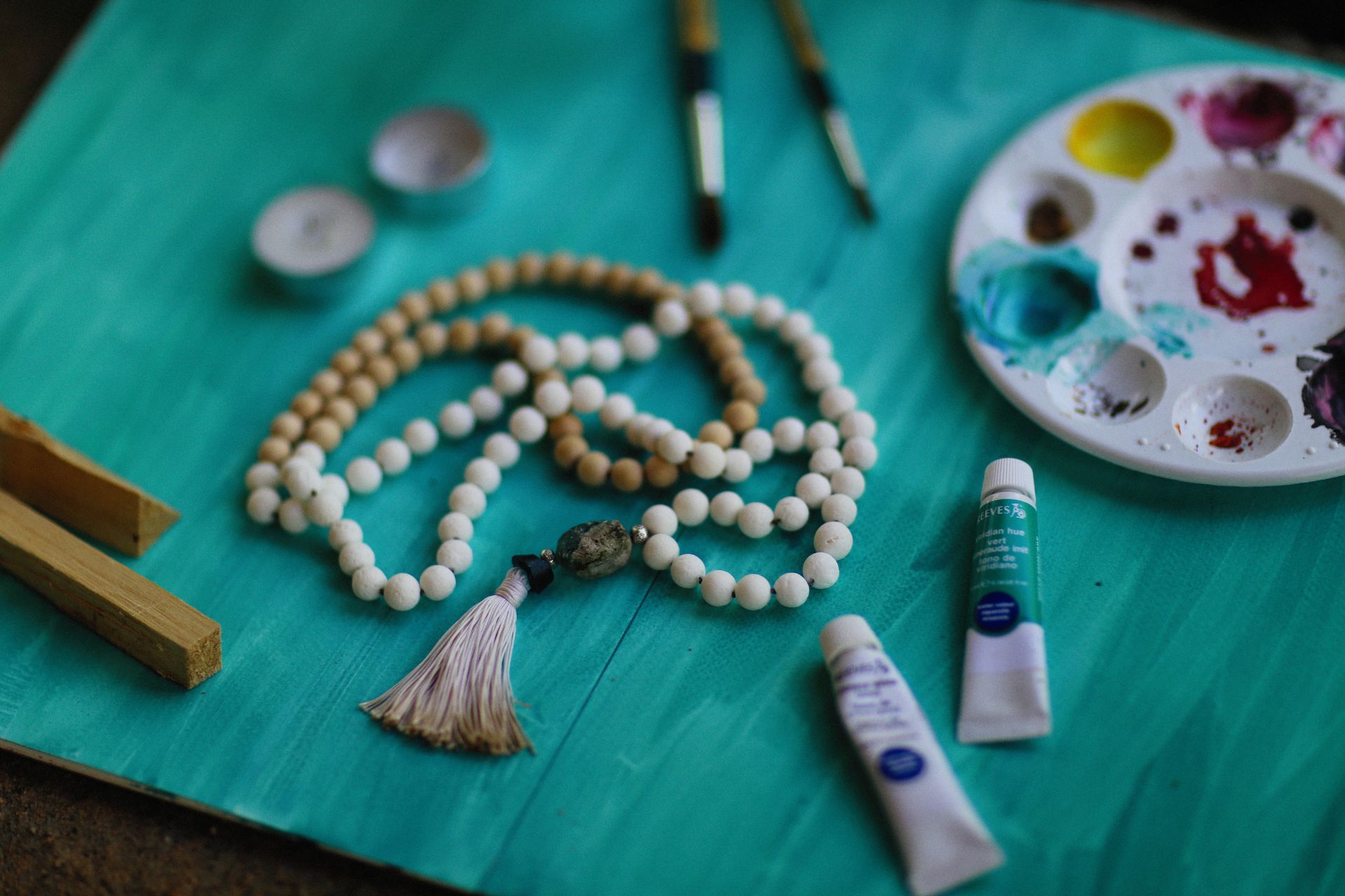 Flat lay of handmade bead necklace with paint and brushes on a turquoise background.