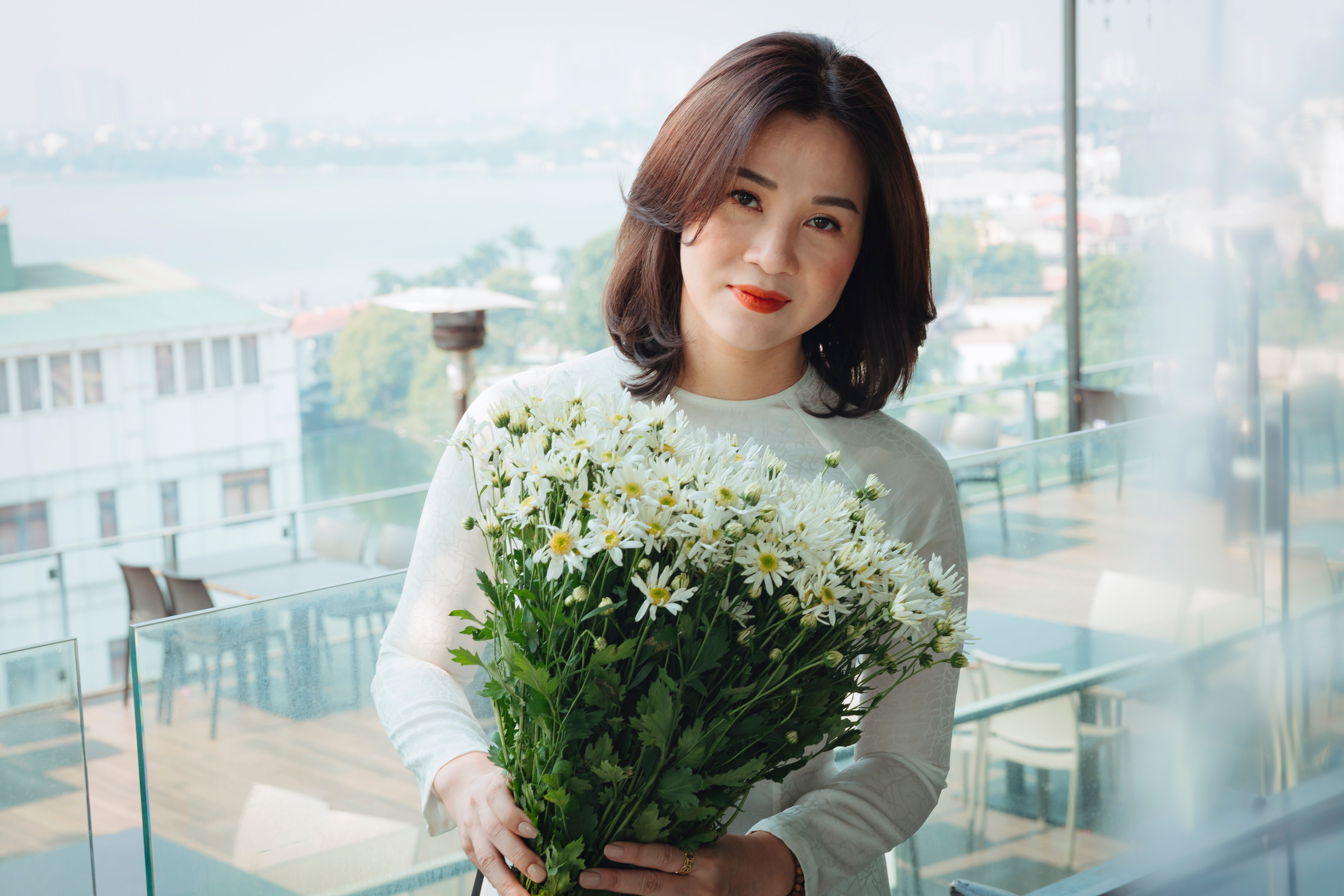 A portrait of a smiling woman holding white daisies on a sunny balcony with a cityscape view.
