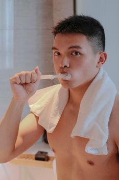 A young man in the bathroom with a towel brushing his teeth, promoting oral hygiene.