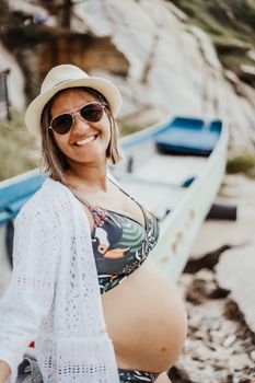 Smiling pregnant woman in sunglasses and hat at the beach, embracing motherhood.