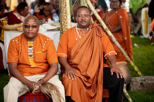 Two men wearing traditional orange attire at a cultural celebration, seated with a festive backdrop.