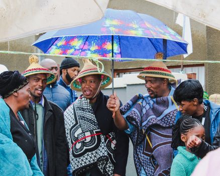 People wearing traditional attire celebrate with a colorful umbrella on a rainy day.
