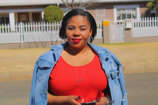 African woman in a red dress and denim jacket confidently holding her smartphone outdoors.