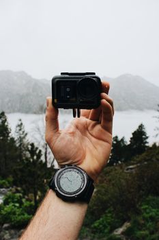 Close-up of a hand holding an action camera with a digital watch in a misty mountain setting.