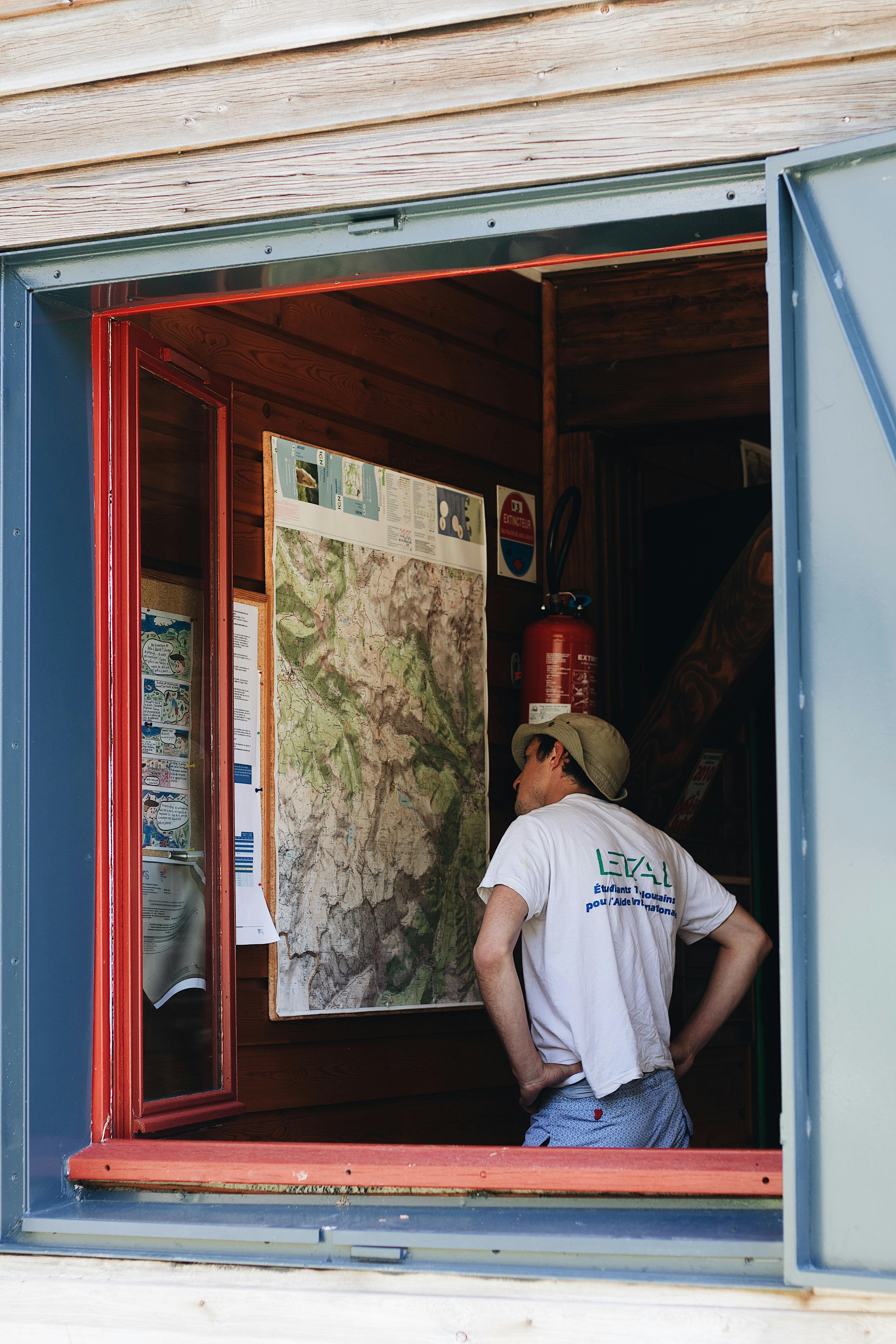 Man wearing cap examining a large map in a rustic room through an open window.