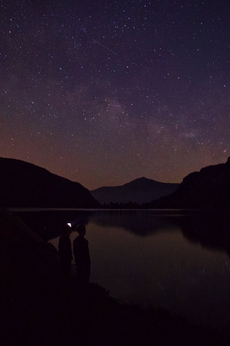 Silhouettes Of People Standing On Lake Shore Under Starry Sky