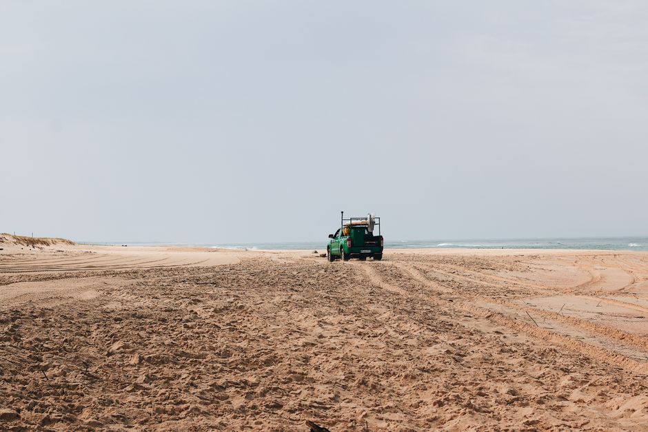 A green pickup truck driving on a vast, sandy beach with ocean waves in the background.