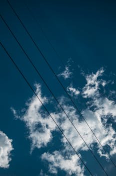 Low angle view of electric wires against a blue sky with white clouds, creating a sense of connection and energy.