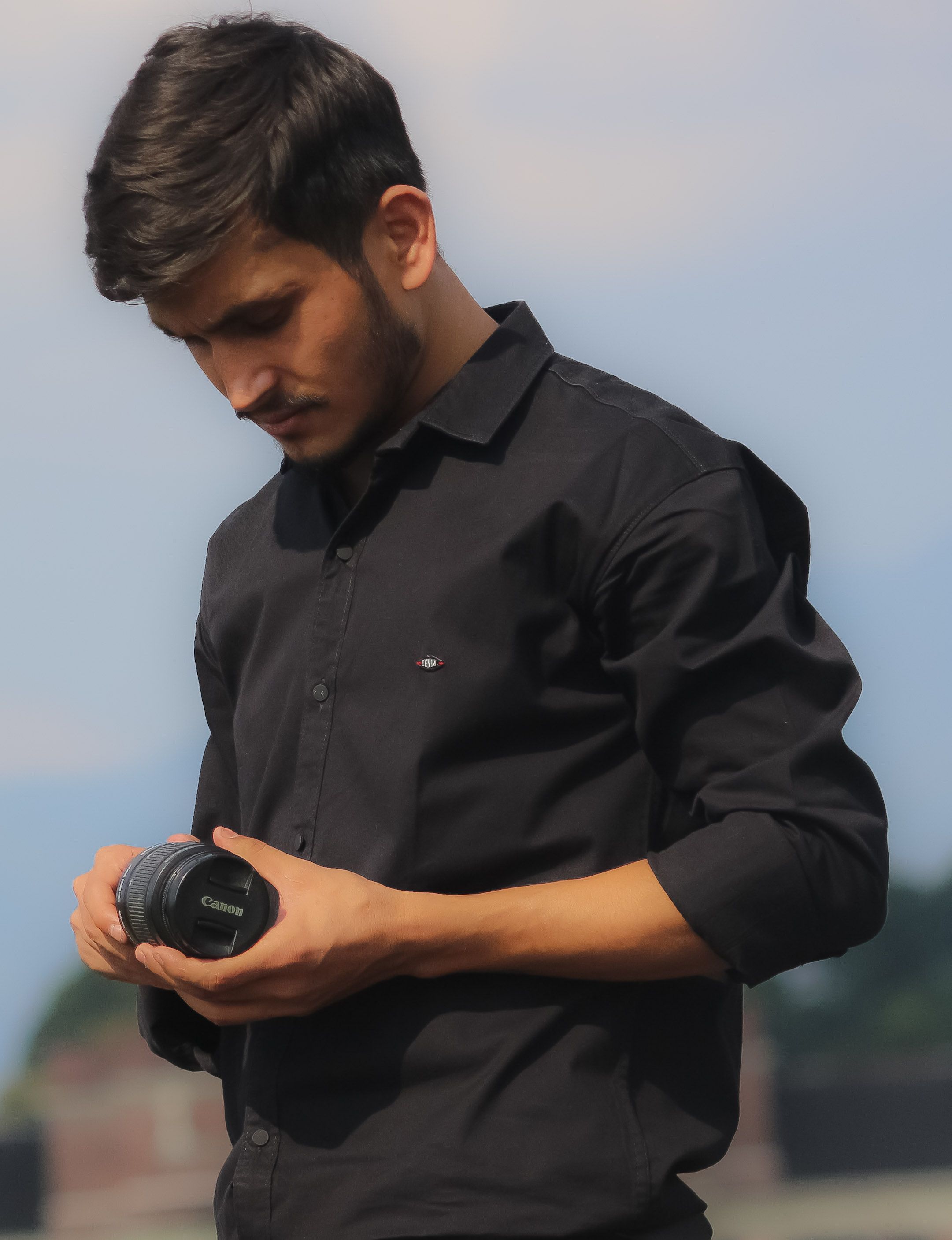 Young man in black shirt inspecting a camera lens outdoors on a sunny day.