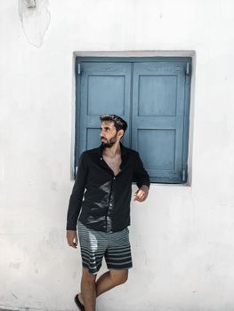 A stylish man in casual attire poses against a white wall with a blue window, exuding charm and confidence.