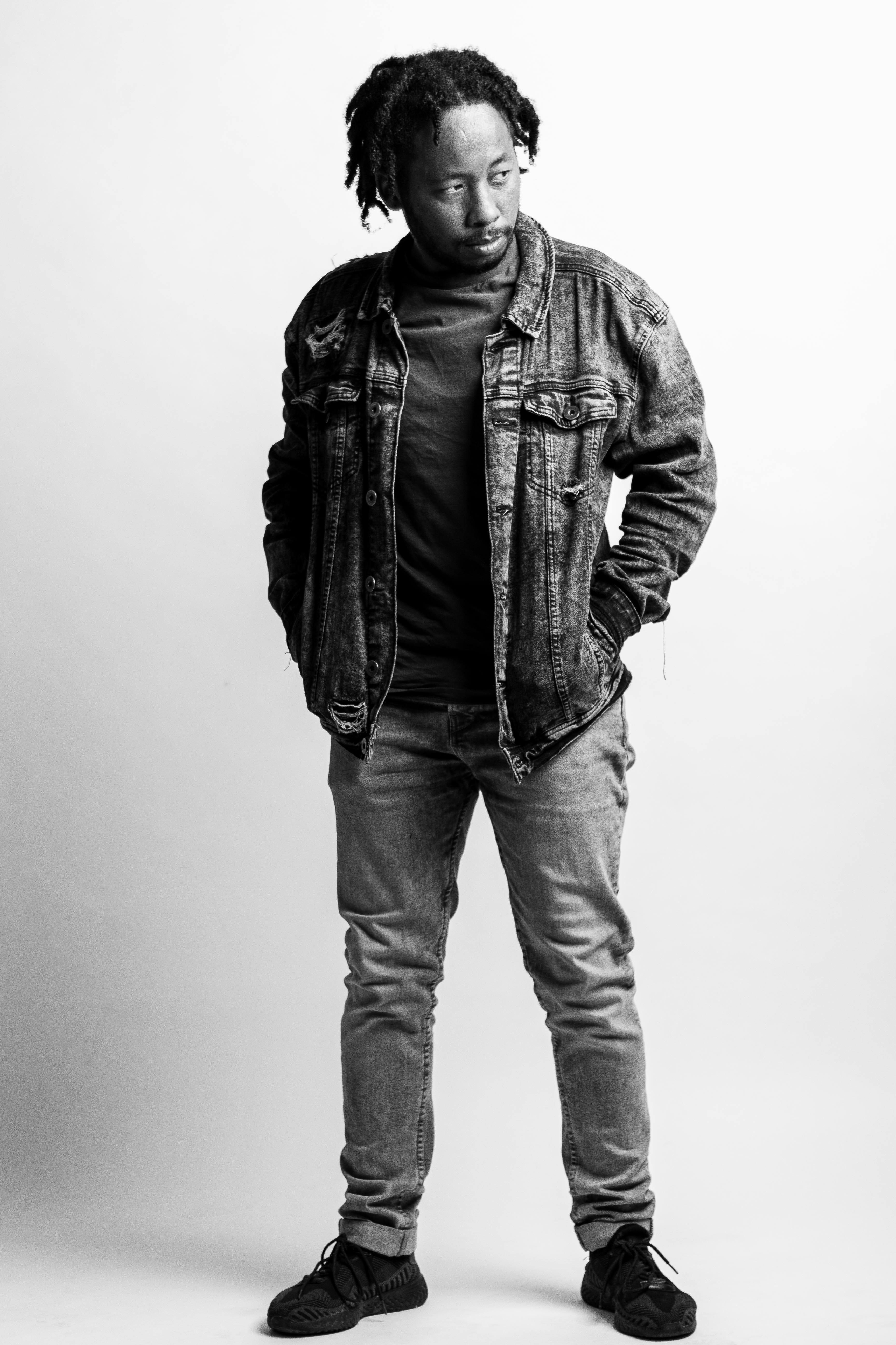 Black and white portrait of a stylish man in a denim jacket standing confidently on a white background.