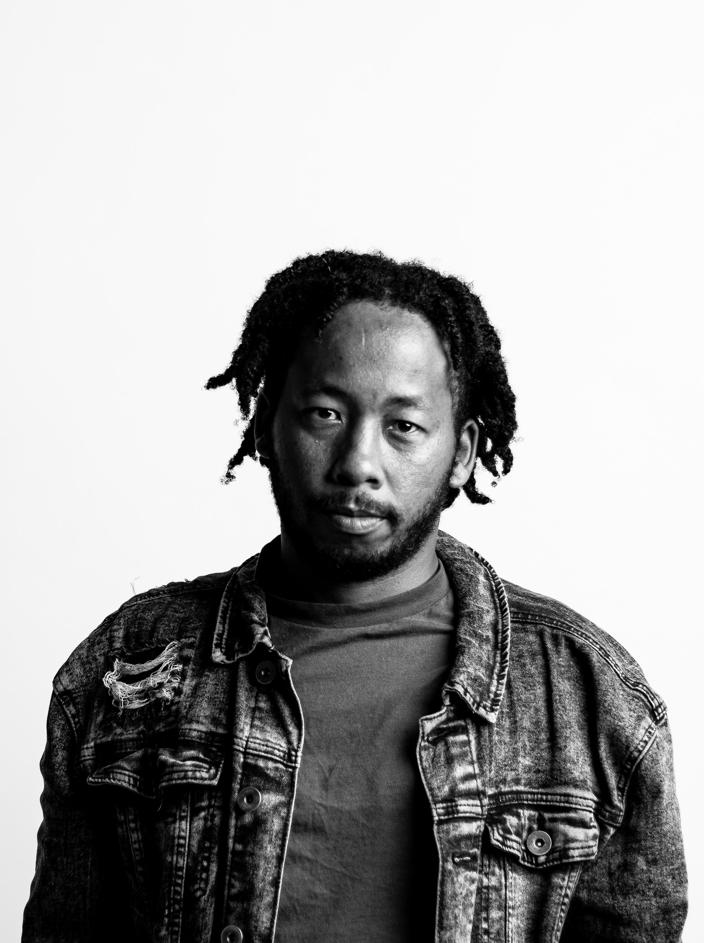 Monochrome portrait of a man with dreadlocks wearing a denim jacket in a studio.