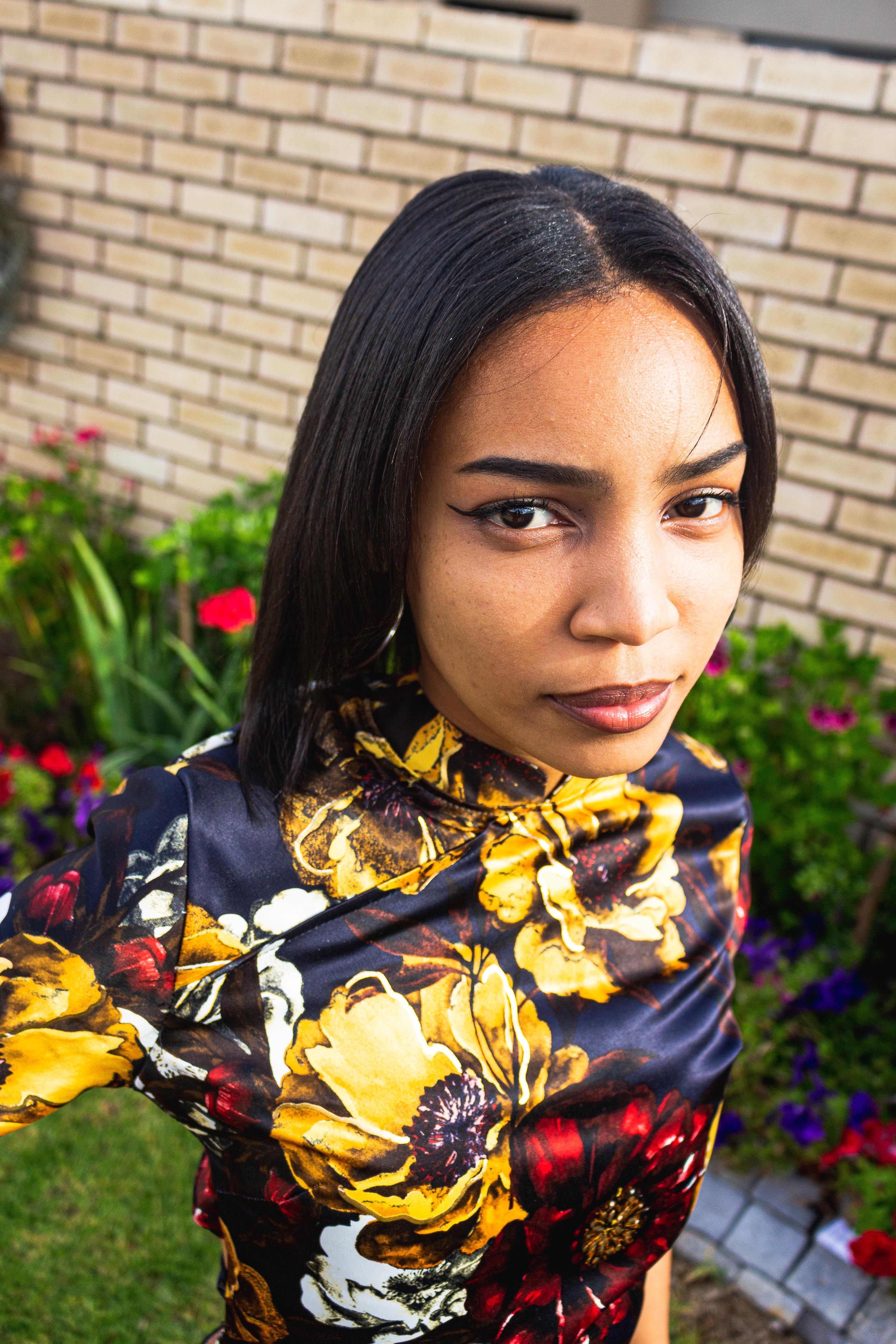 A young woman wearing a floral dress poses confidently against a brick wall.