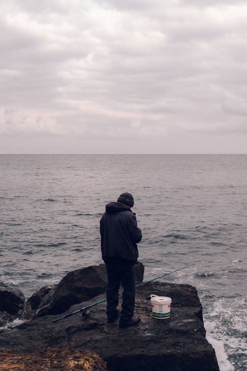 Free Solitary fisherman stands on rocky shore of Türkiye under a gloomy sky, casting into the sea. Stock Photo