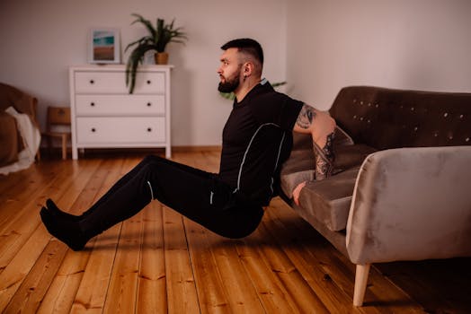 A bearded man in black fitness attire exercising at home, showcasing arm tattoos and a healthy lifestyle.