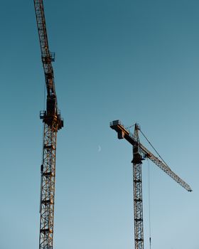 Two construction cranes silhouetted against a clear blue sky during daylight.