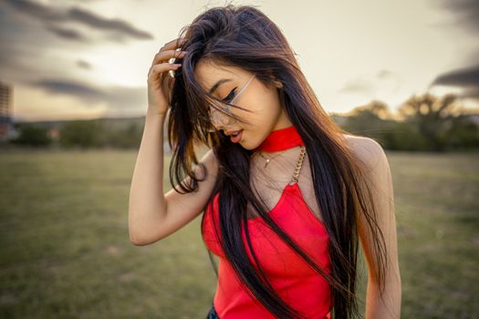 A fashionable woman in a red top and eyeglasses posing in a serene outdoor setting during sunset.