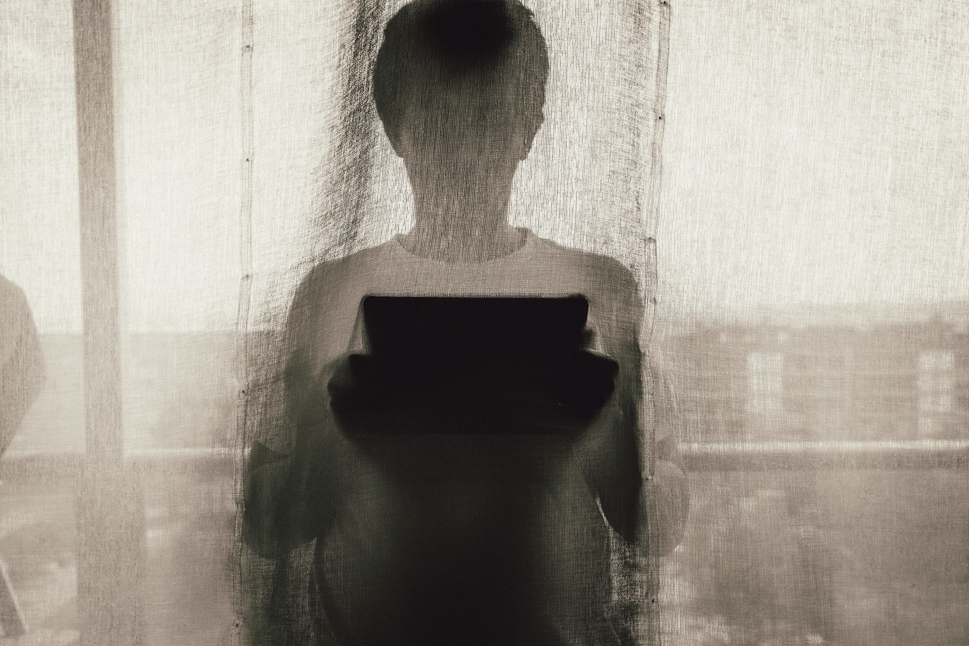 A silhouette of a person using a tablet device behind a curtain indoors, creating a mysterious ambiance.