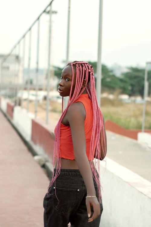 Free A fashionable woman with long pink braided hair looks back while walking outdoors in a vibrant red crop top. Stock Photo
