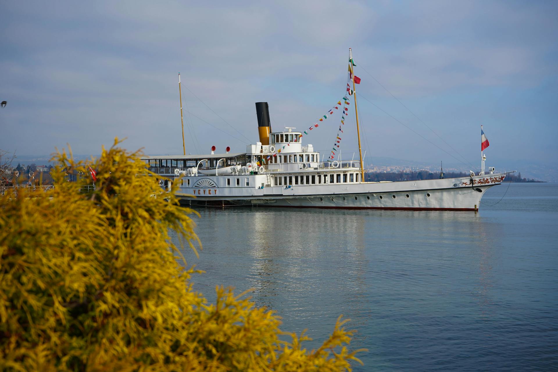 A classic paddle steamer docked by a scenic lake with flags and clear skies.