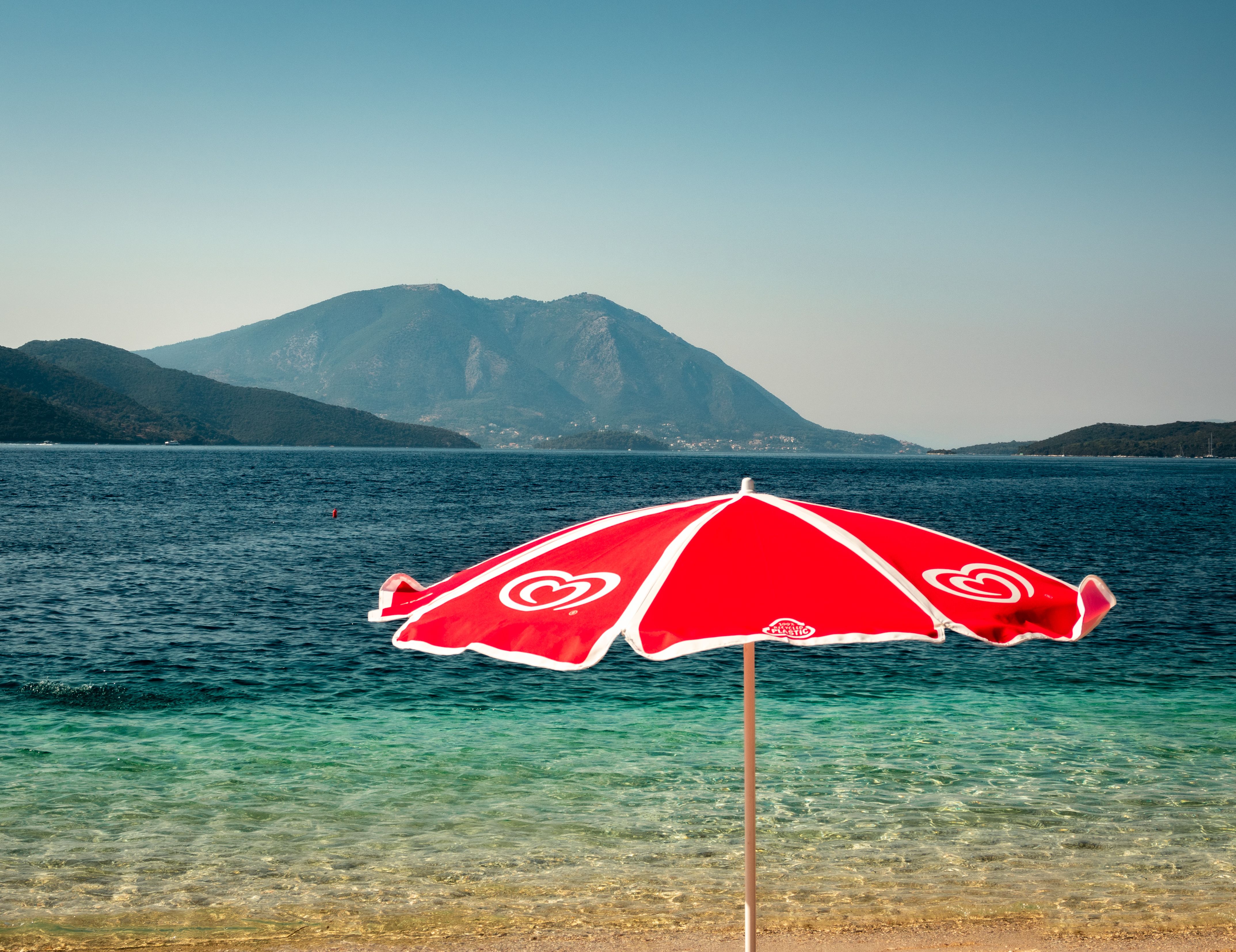 Red beach umbrella with serene sea view and distant mountains in Greece.