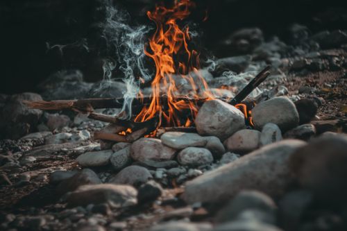 Free Warm campfire surrounded by stones with orange flames and smoke rising in a dark outdoor setting. Stock Photo