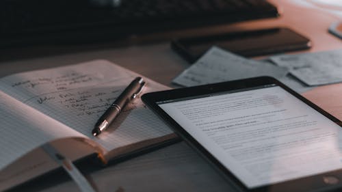 Close-Up Shot of a Tablet beside a Notebook and a Pen