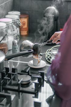 A woman pours steaming Turkish coffee into a cup on a modern kitchen stove.