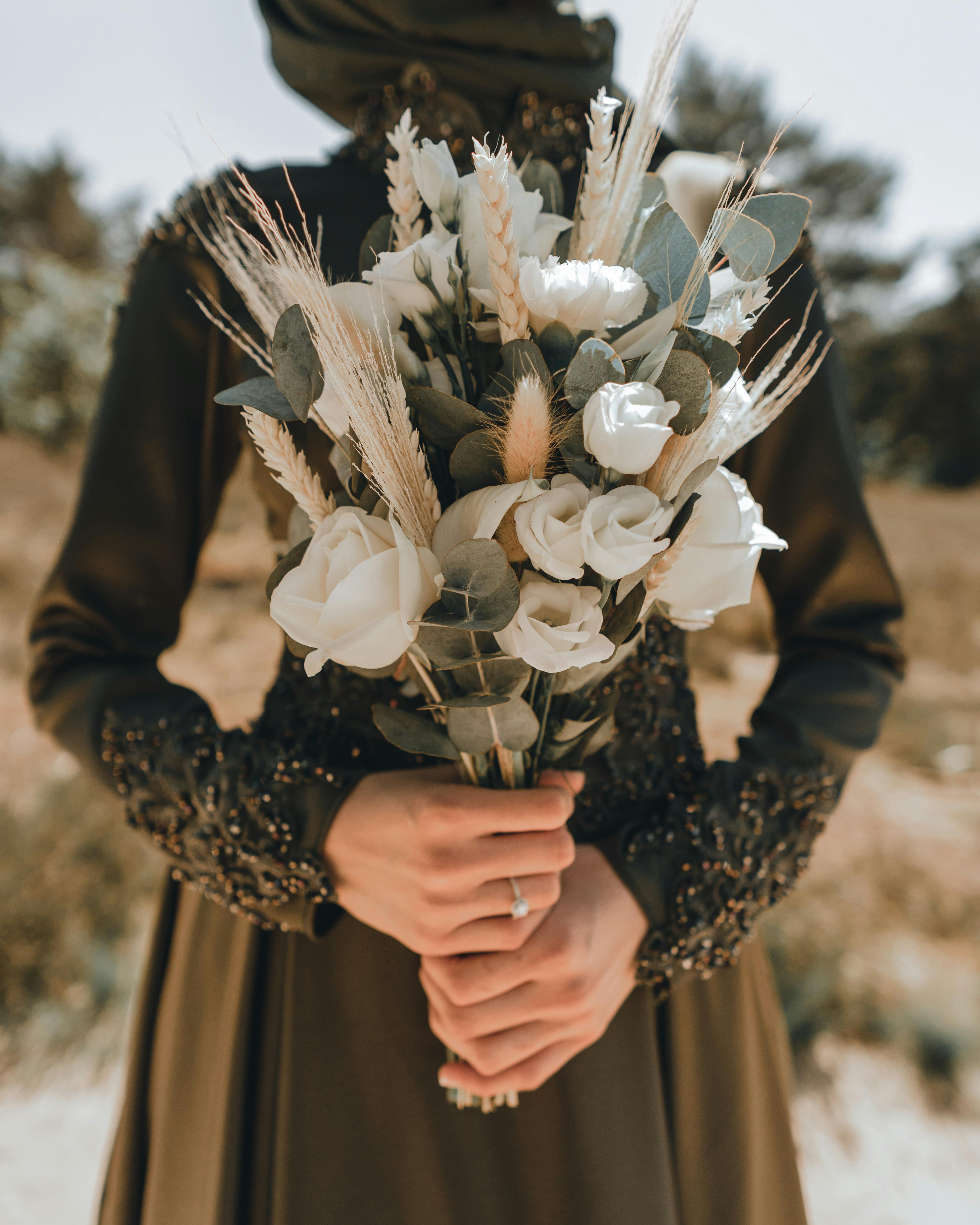 A woman in a formal dress holding a bouquet of white flowers with greenery outdoors.